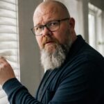 Thor Jadesø kigger ud gennem persiennerne i sit kontor og ser mistænksomt direkte ind i kameraet, som om nogen holder øje med ham