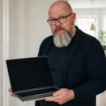 Thor Jadesø står i en lys entré og holder en åben bærbar frem mod kameraet med løftede øjenbryn og en advarende mine