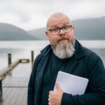 Thor Jadesø står på en træbrygge ved en grå norsk fjord med et papir i hånden og kigger overrasket direkte i kameraet